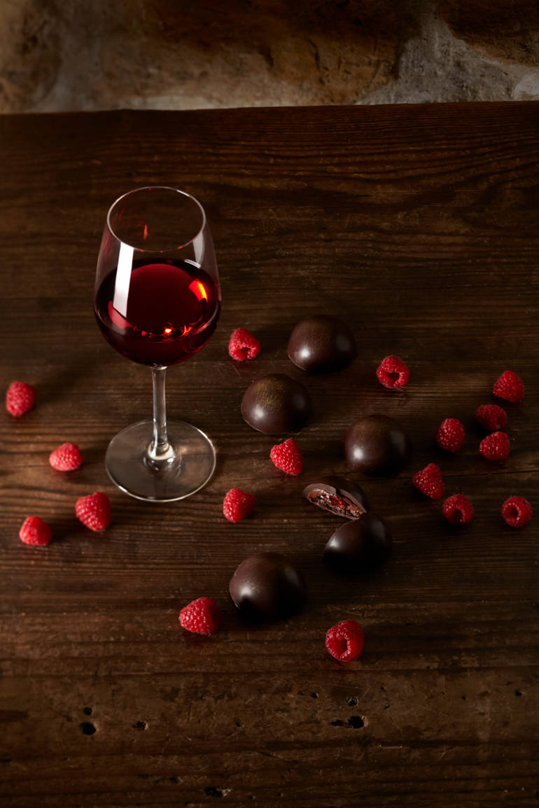 Un verre de vin rouge est posé sur une table en bois entouré de framboises et de bonbons au chocolat.