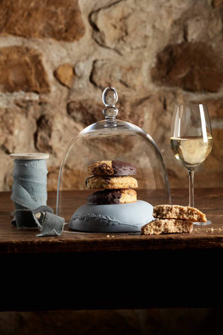 Une cloche en verre recouvre des biscuits, accompagnée d'un verre de vin blanc et d'une bobine de fil sur une table en bois.
