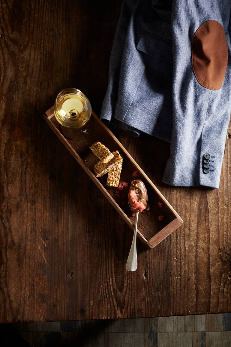 Une assiette en bois contient des morceaux de nourriture et un verre de boisson, avec une veste en arrière-plan sur une table en bois.