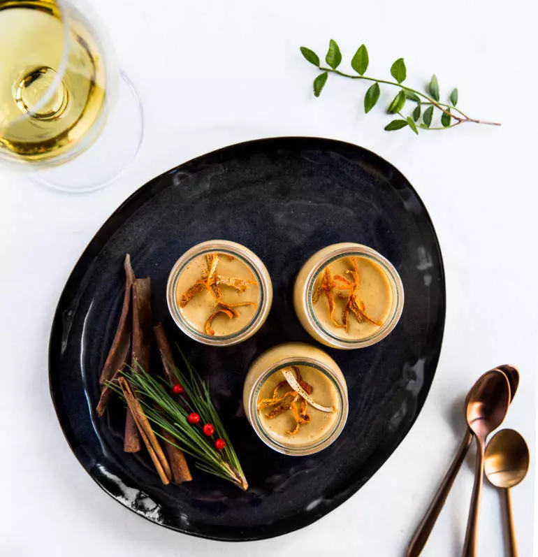 Trois petits pots de crème garnis de zeste, accompagnés de bâtons de cannelle, d'aiguilles de pin et d'un verre de vin blanc, le tout présenté sur une assiette noire.
