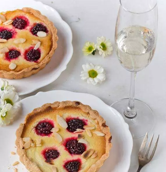 Deux tartelettes aux mûres garnies d'amandes effilées, accompagnées de fleurs blanches et d'un verre de champagne.