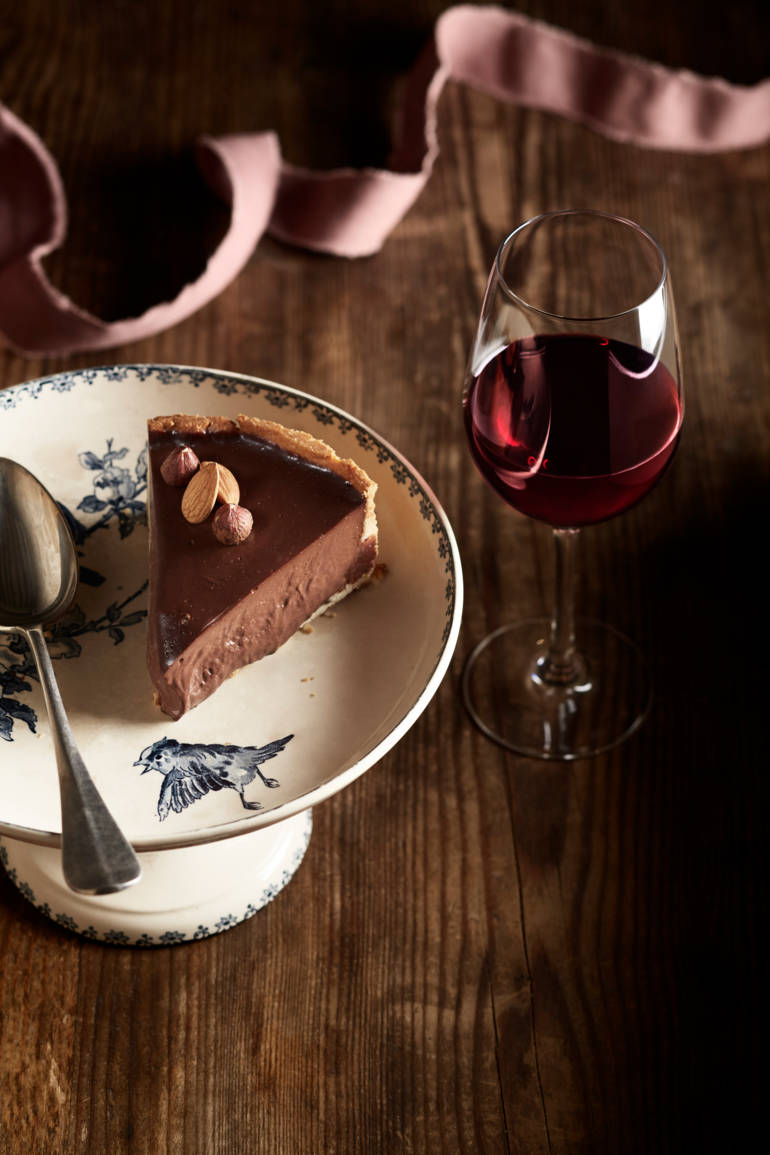 Une part de gâteau au chocolat décorée d'amandes et de noisettes, servie sur une assiette ornée, accompagnée d'un verre de vin rouge.